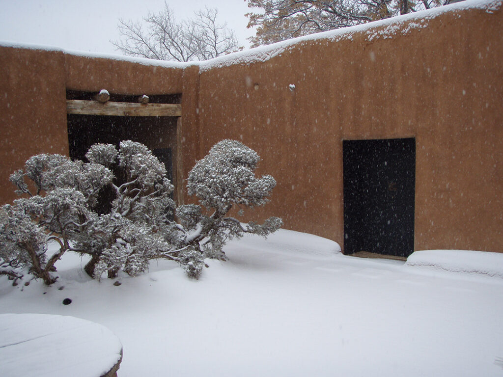 Photograph of an interior courtyard with a dark door on the right and square opening on the left with beams above. In the courtyard is the top of a circular well and a small trees. The entire space is covered in white snow.
