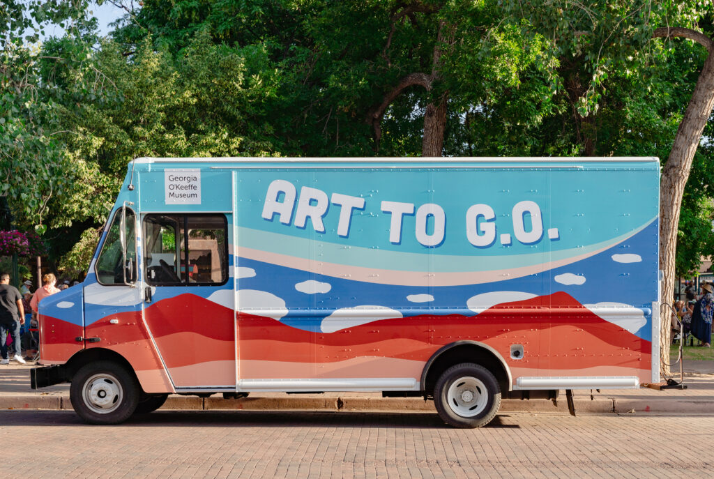 Photograph of a truck with a desert horizon and cloud motifs on the side along with the heading 'Art to G.O.' and the Georgia O'Keeffe Museum Logo. Behind the truck are crowd of people under green trees.