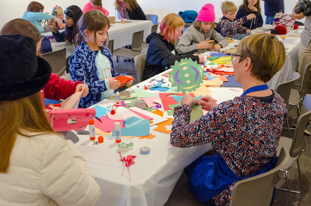 Photograph of a group of children and adults all sitting around along table full or papers and art supplies. They are all concentrated on making crafts.