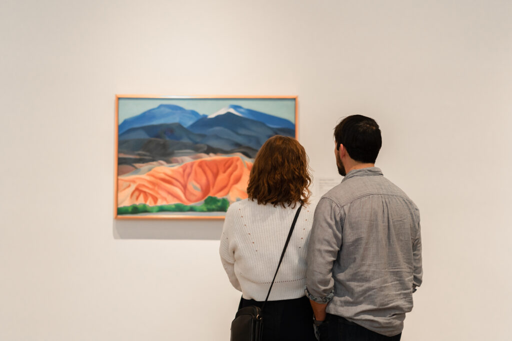 A photograph of two people taken from behind, they are standing close together looking at O’Keeffe’s painting ‘Black Mesa Landscape, New Mexico / Out Back of Marie’s II’ in the Museum galleries.