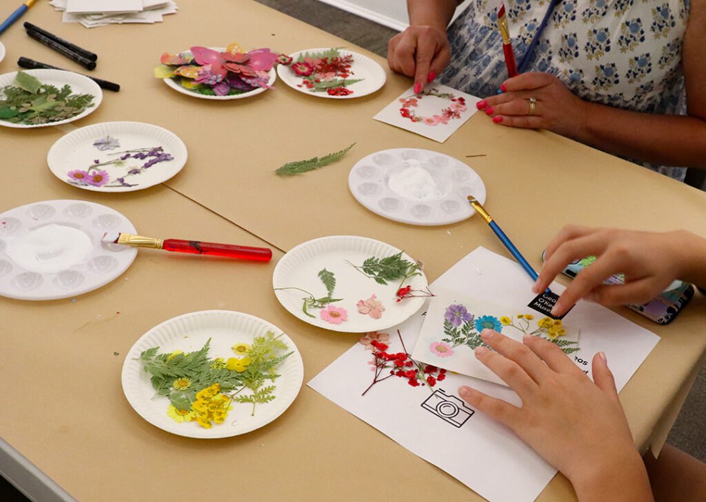 Photograph of a table with art supplies and small leaves and flowers. Two people are sitting on the edges of the tables their hands visible in the photograph. They use their fingers and tools to press little flowers and leaves into paper.