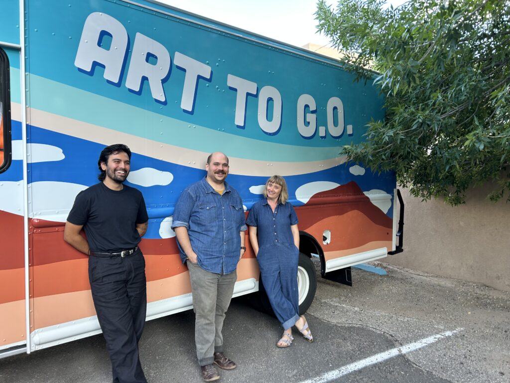 Three people smiling and standing in front of the Art to G.O. Art Truck with clouds motifs on it. On the right is the edge of a leafy green tree.