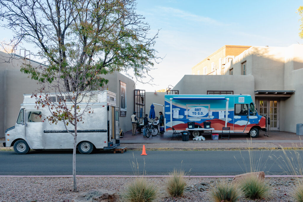 Photograph of a street and an adobe building with a tree on the left of the frame. Parked in front of the building and on the street are two trucks, one a grey mobile art gallery and one the Art to G.O. truck with cloud motifs on it and two tables in front.