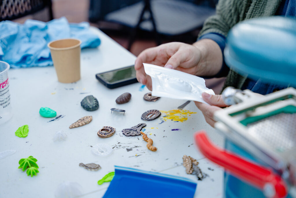 Close-up photograph of a pair of hands holding a plastic mold over a table. On the table are small colorful plastic creations.