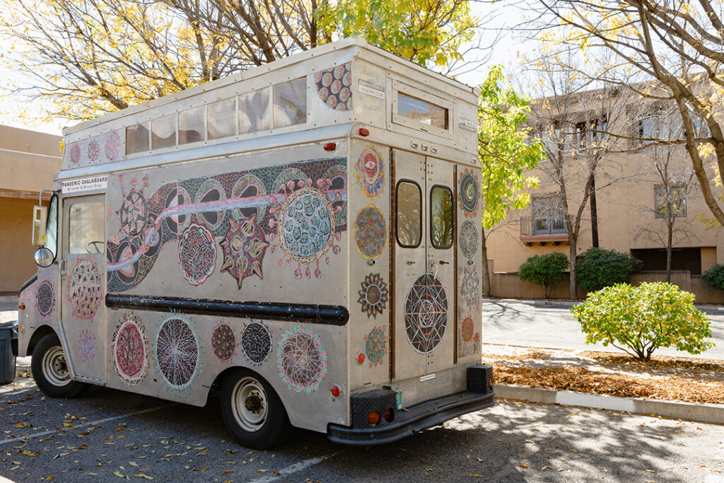 Photograph of a rectangular grey truck, a mobile art gallery, with artwork on the sides and front. The truck is parked underneath leaves with golden foliage.