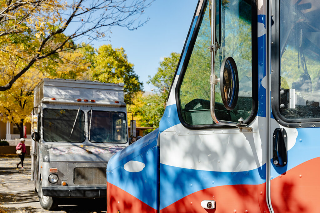 Photograph of the front of a grey mobile art gallery in the background and the front of the Art to G.O. truck in the foreground. Behind both trucks are leafy trees turning gold.