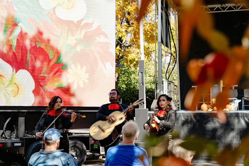 Three people in dark clothes holding and playing instruments including a guitar and a violin. Behind the is a large screen with a red paintings by O'Keeffe and surrounding the scene is fall folliage