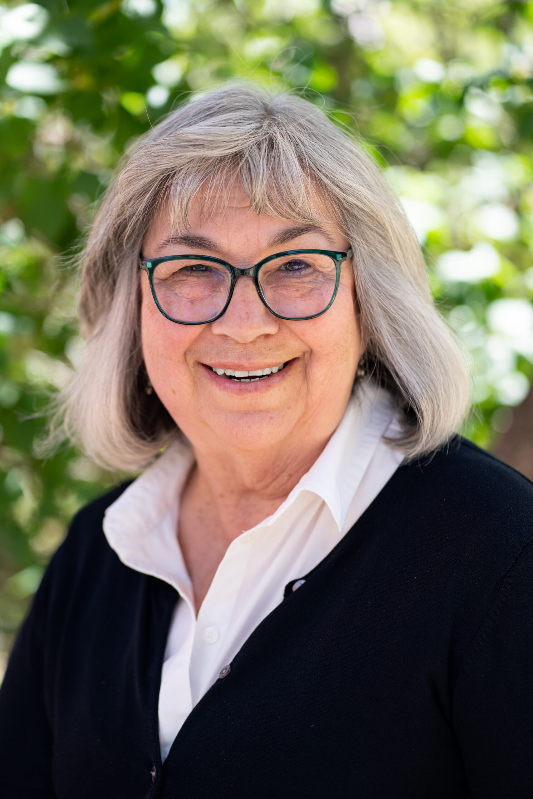Headshot of a person with short hair and glasses smiling at the camera with green foliage behind.