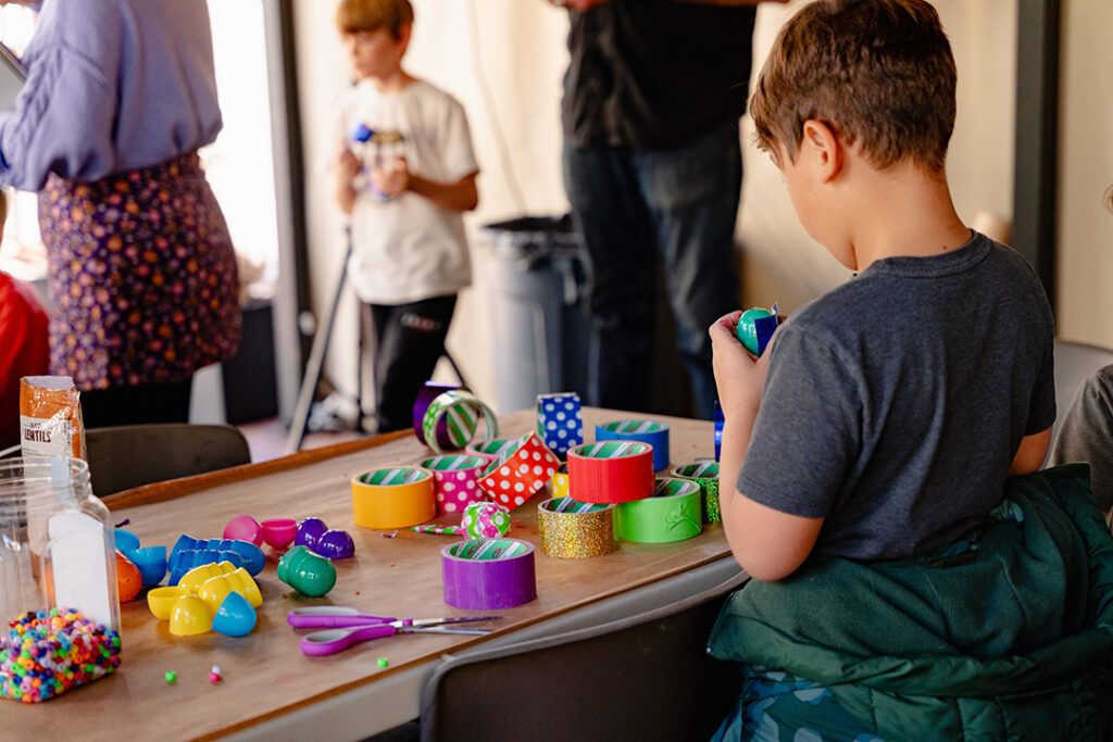 A young child at a table covered in colorful rolls of tape. The photo is taken from behind as the child looks down at the rolls. In the background are a few adults and other children.