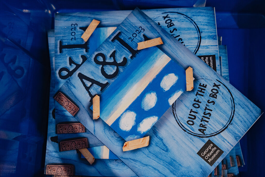 Photograph of a pile of blue booklets with an illustrated cover and the text "A&L 2023, Out of the Artist's Box"