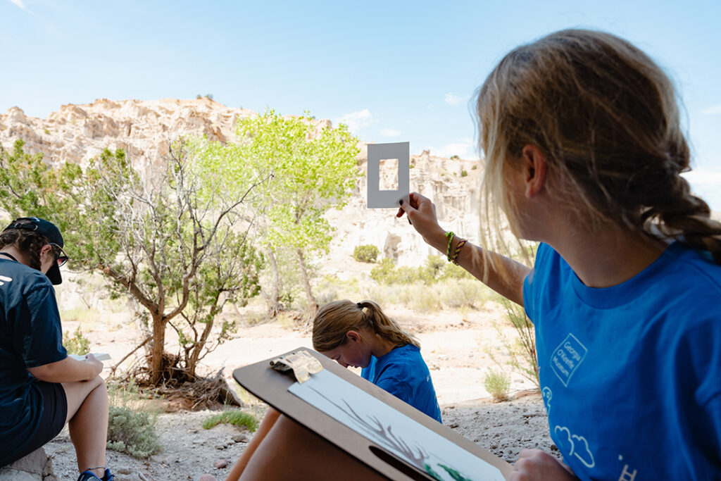 Photograph of three young people outside in the desert all sketching on notepads. On the right of the frame a person is holding up a small frame of paper as a reference for their drawing. In the background are  desert trees and a blue sky.
