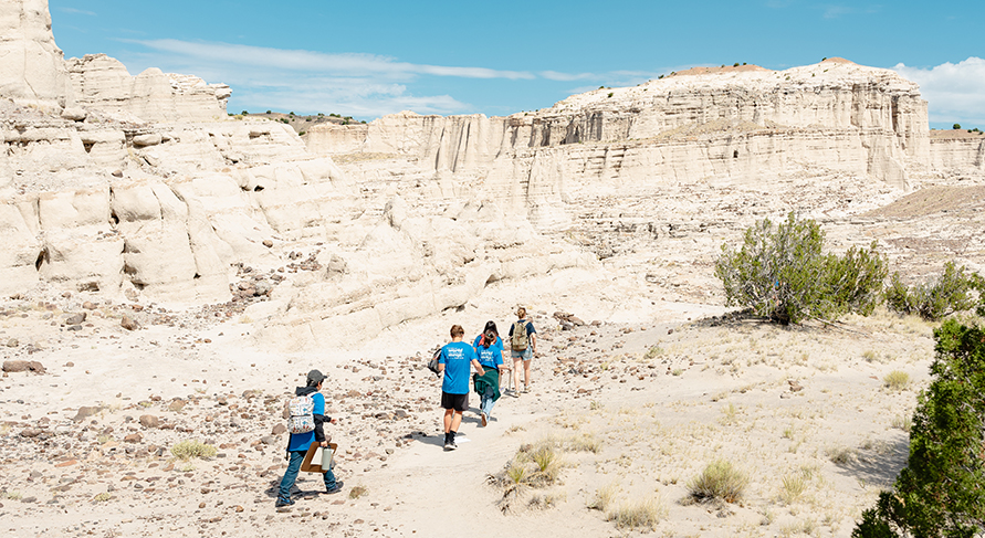 Photograph of a group of young students, many wearing blue shirts, backpacks, and hats, walking through the desert landscape known as the Plaza Blanca, or the White Place. They are small in the frame and the desert landscape extends all around them. The upper portion of the frame shoes a clear blue sky.