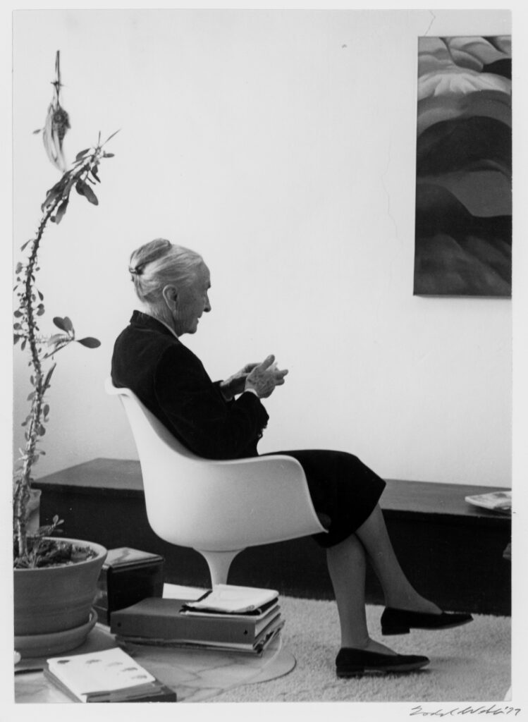 Full length profile view of O'Keeffe sitting in a white modular chair in her studio, her hair is pulled back into a bun, she wears a black suit coat and skirt, nylons, and black flat leather designer shoes while holding an object in her hand, hanging on the wall in viewer right is portion of one of her paintings (Black Place?), behind her to the left on a glass table are binders, books and a large spindly plant