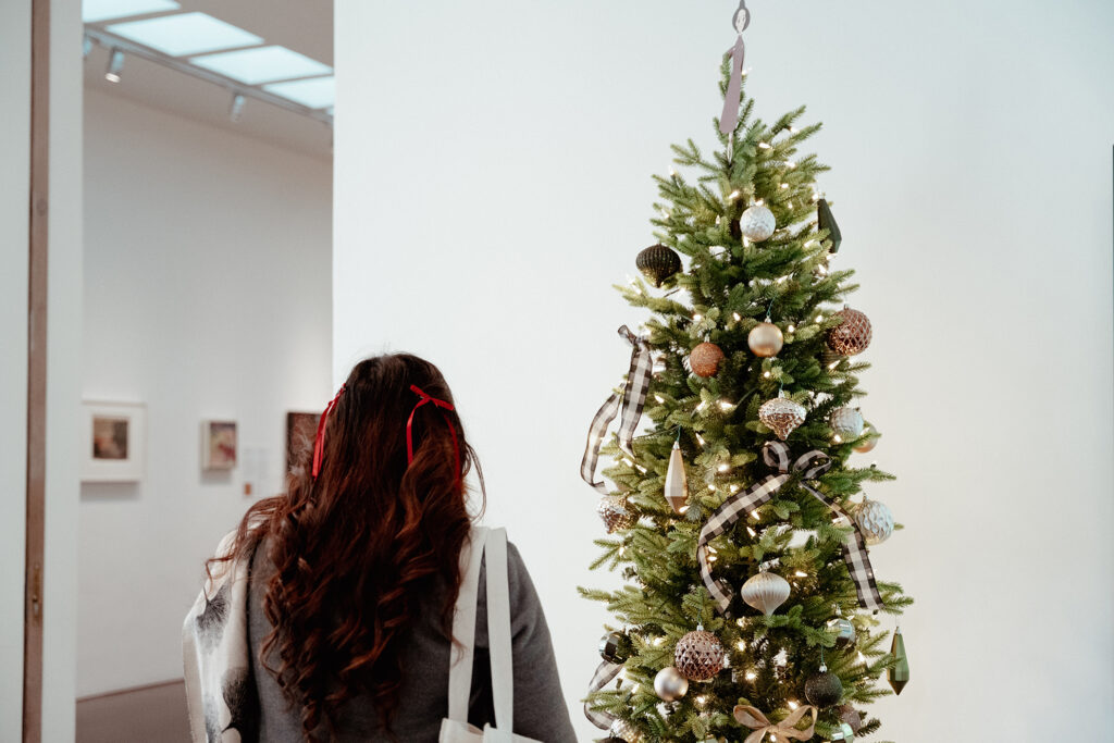 A person with long dark hair and small red bows in their hair standing to the left of a christmas tree. In front of them is an opening into a Museum gallery.