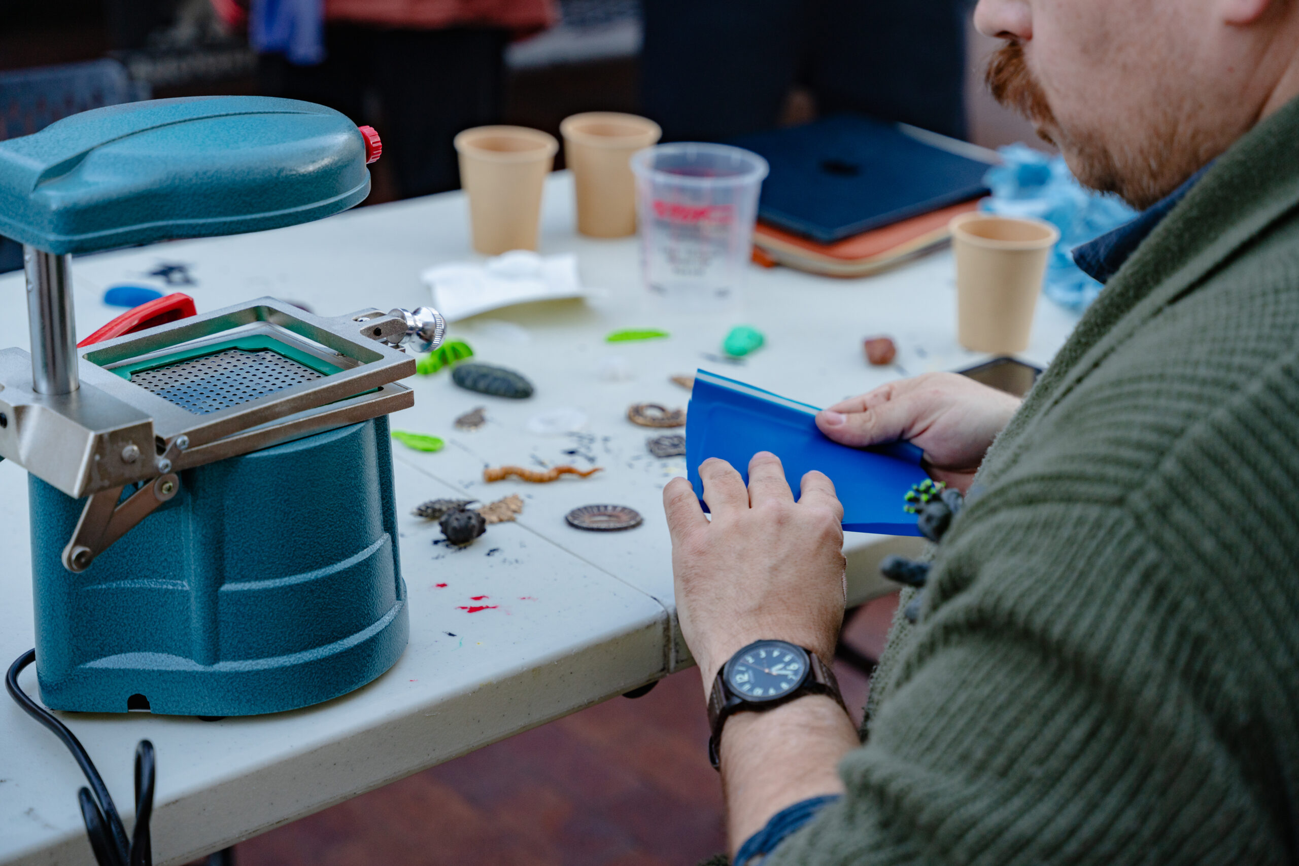 A photograph of a table with industrial style art materials. In the right hand corner of the photograph a man holds art supplies ready to use the machine in the left corner of the table.