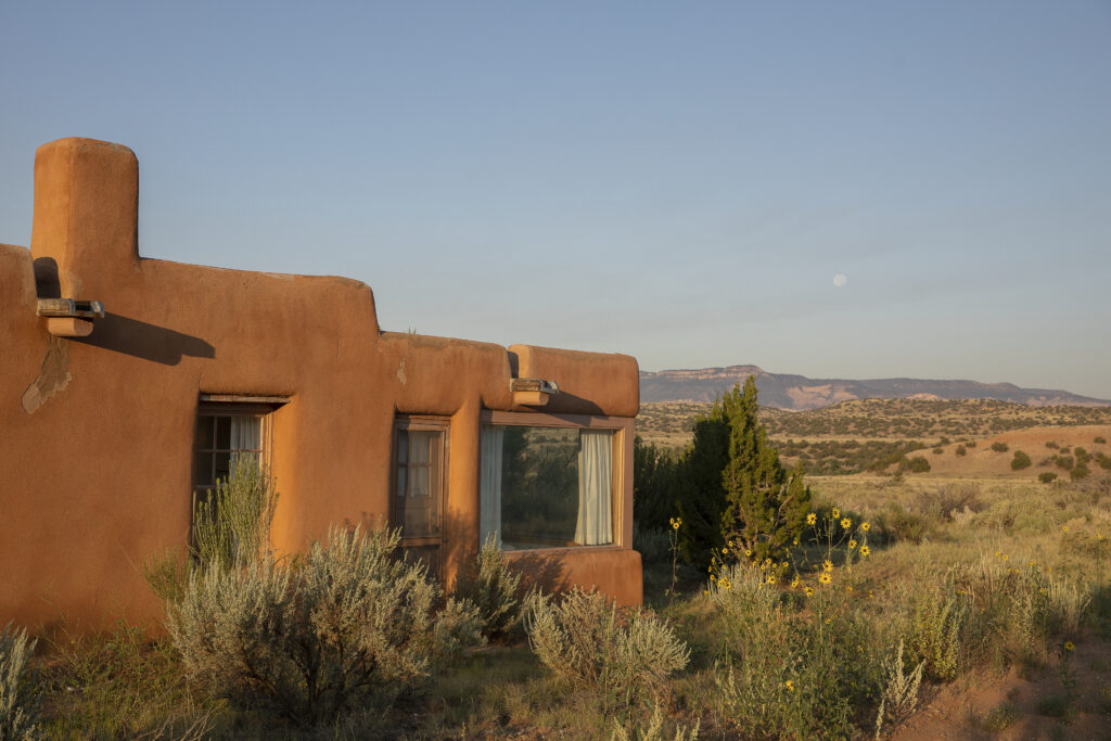 Photograph of the exterior of an adobe home with several windows. Behind the home is a mountain across the horizon and a clear blue sky.