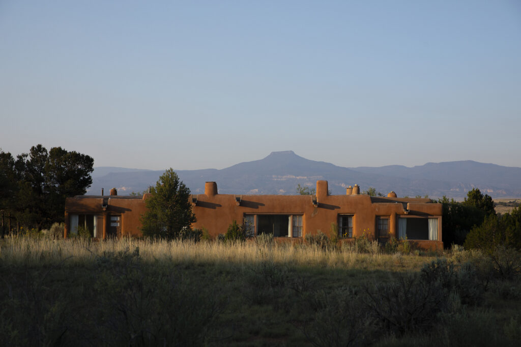 Photograph of the exterior of an adobe home with several windows. Behind the home is a mountain across the horizon and a clear blue sky.