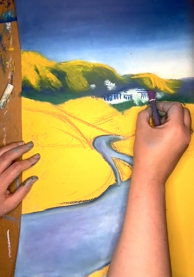 The view of a right hand using pastels to create a landscape with a yellow grasses and a winding river flowing through it.