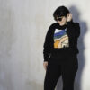 Person with dark hair and sunglasses wearing a black sweatshirt with a simple modern design of a desert landscape and adobe home in shades of blue, beige, green, and orange.