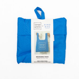 A reusable bag in blue with the Georgia O'Keeffe Museum Logo on it. It is folded into a small square.
