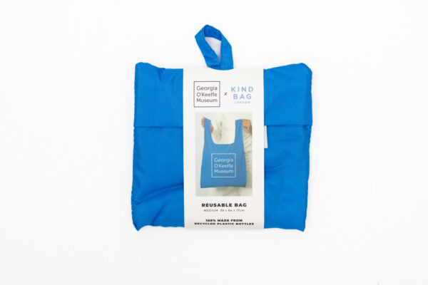 A reusable bag in blue with the Georgia O'Keeffe Museum Logo on it. It is folded into a small square.