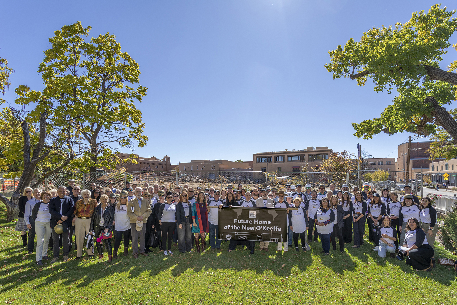 Photograph of a large group of people standing between two trees on a green lawn. They stand behind a banner that says "Future Home of the New O'Keeffe." Behind the group is a construction site. A blue sky in the top half.