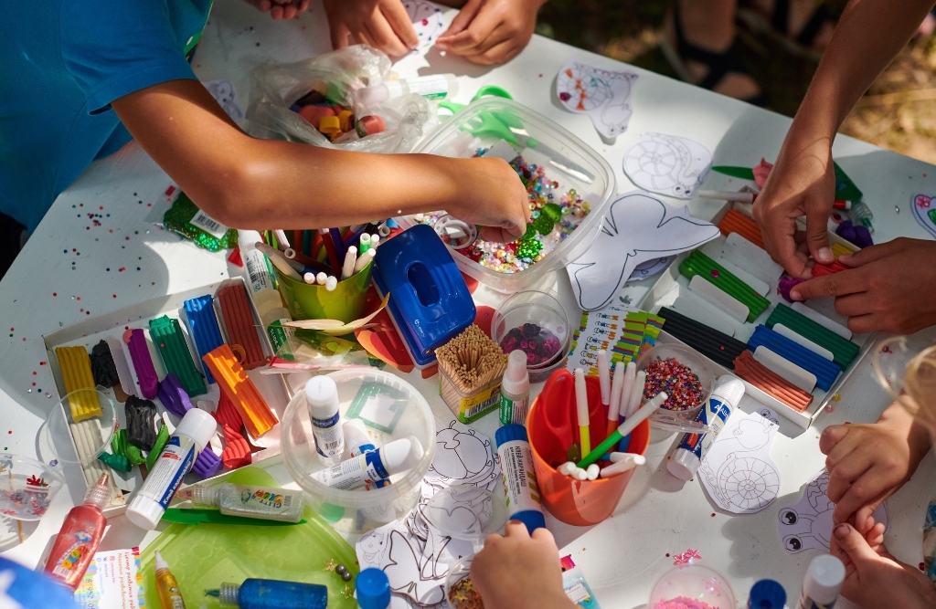 Close-up of colorful art supplies like beads on a table with a group of children's hands using them to make art.