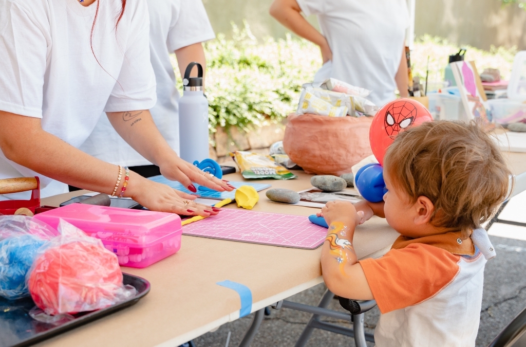 A child sits a table looking at an adult in front of them rolling out a piece of blue clay.
