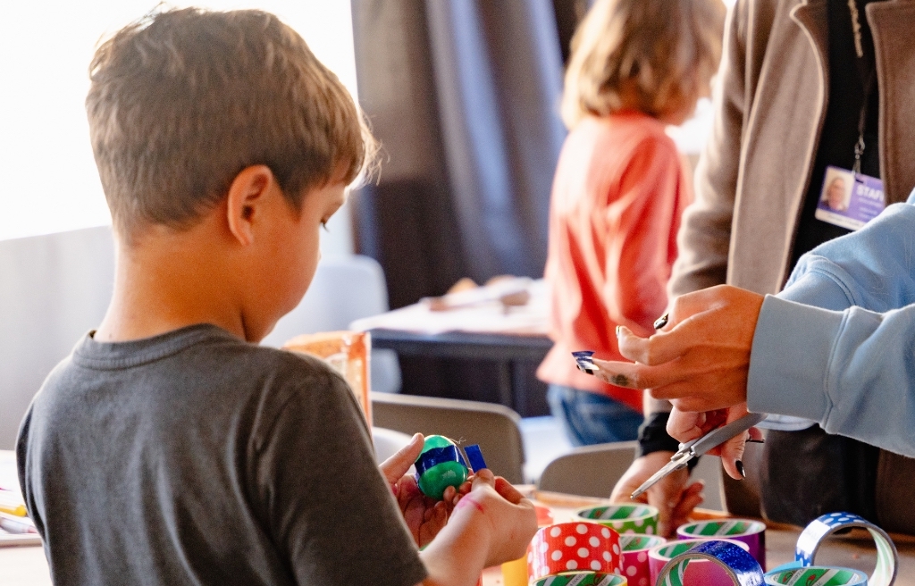 Photograph of a child using colorful tape during an art activities. An adults hands approach from the right side of the frame.