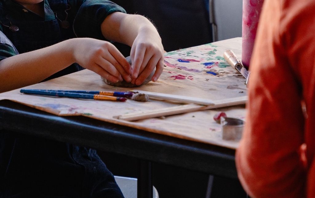 Close-up photograph of a child's hands as they mold clay on a table. On the table are also some paint brushes.