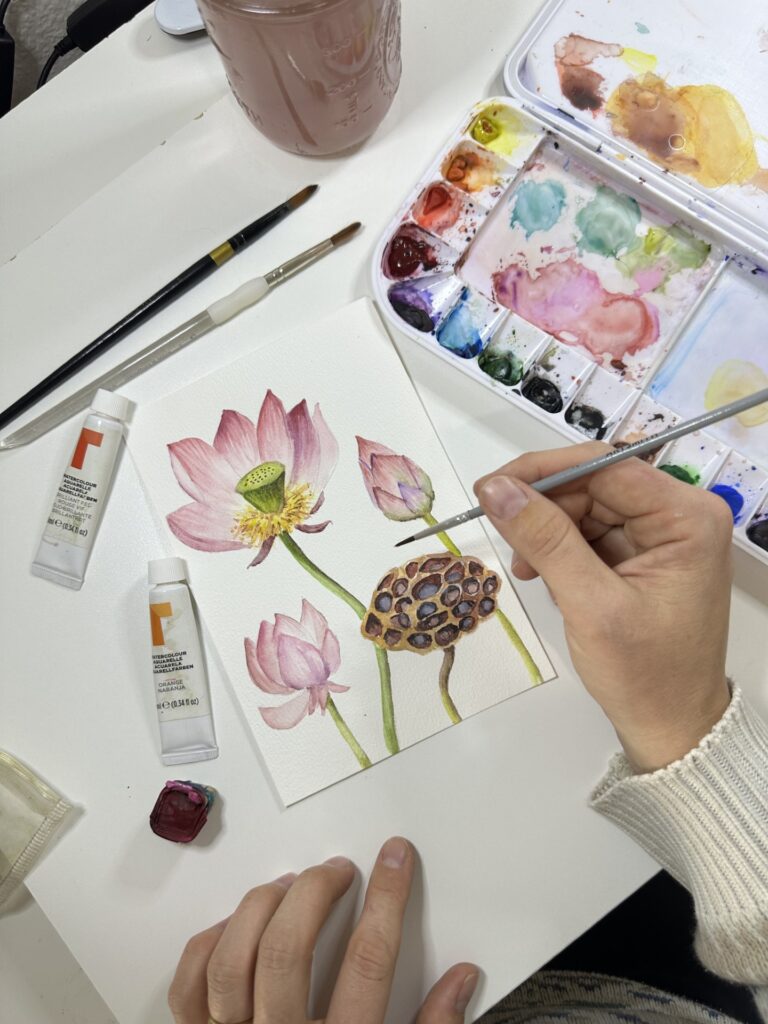 Photograph of an artist's hands from above as they plant several botanical subjects such as a light pink flower. To the right is a watercolor palette.