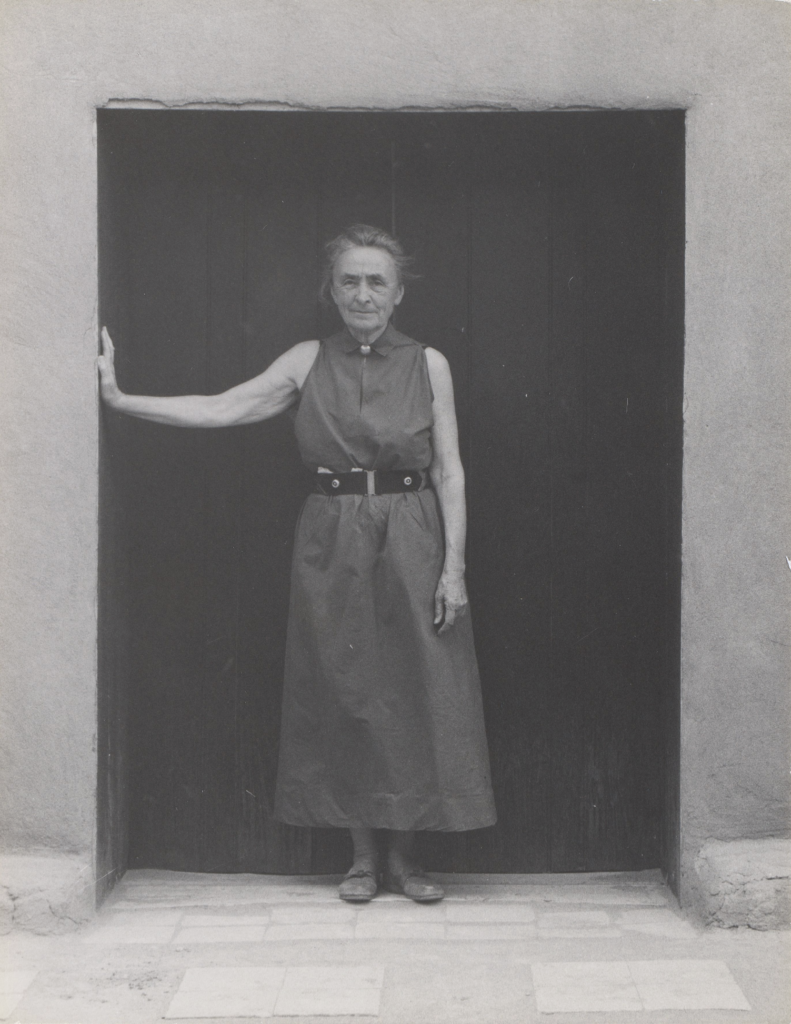 Full length portrait of O'Keeffe standing in the darkened doorway of her Abiquiu house facing the patio; her hair is pulled back from her face, she wears a sleeveless high-collared ankle length dark cotton dress belted with her favorite Taxco black suede and sterling sliver belt; she stands facing the camera with her right arm ( in viewer left) extended out to the wall with her hand leaning on the adobe entrance way.