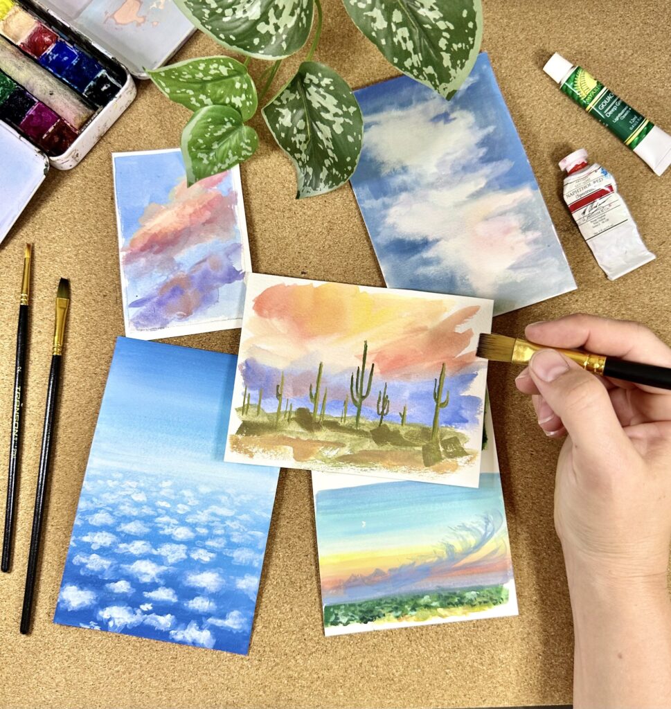 Photograph of an artist's hand as they paint colorful skies on small canvases on a table. Above the paintings is a plant and a few paint tubes.