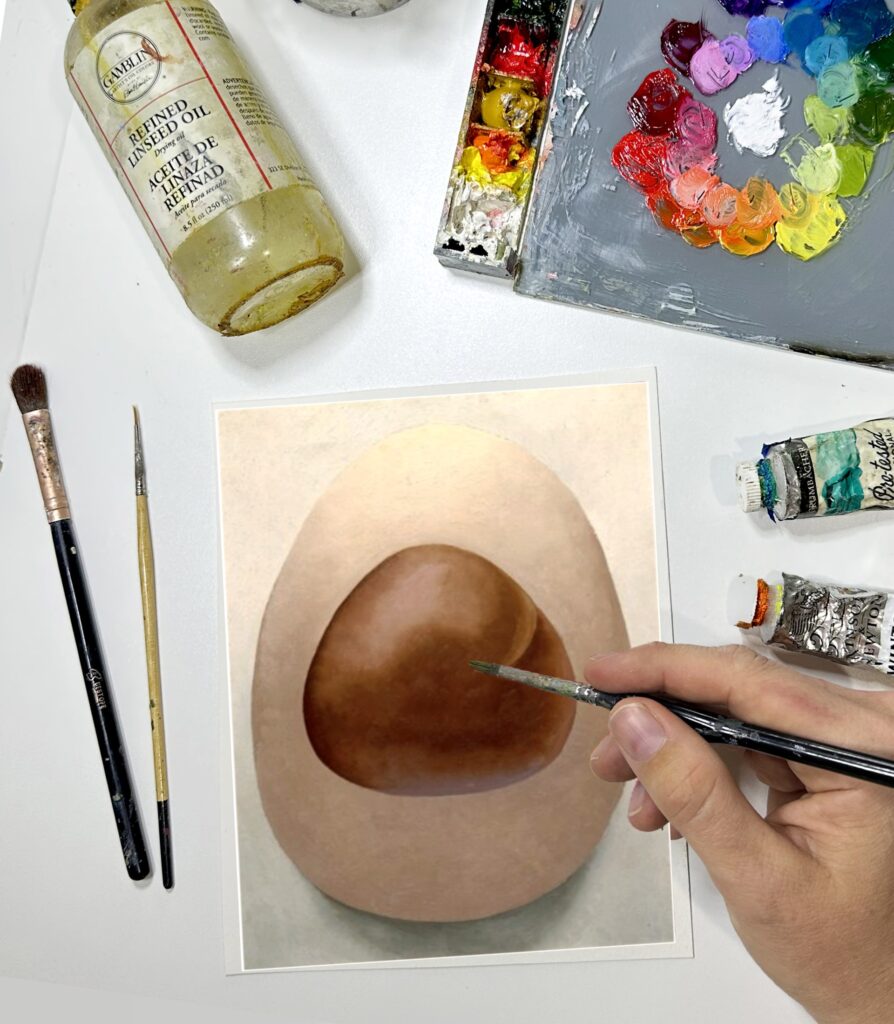 Photograph from above of an artist's hands as they paint a round brown stone on a small piece of paper. Around their hand are paint supplies and a colorful palette.