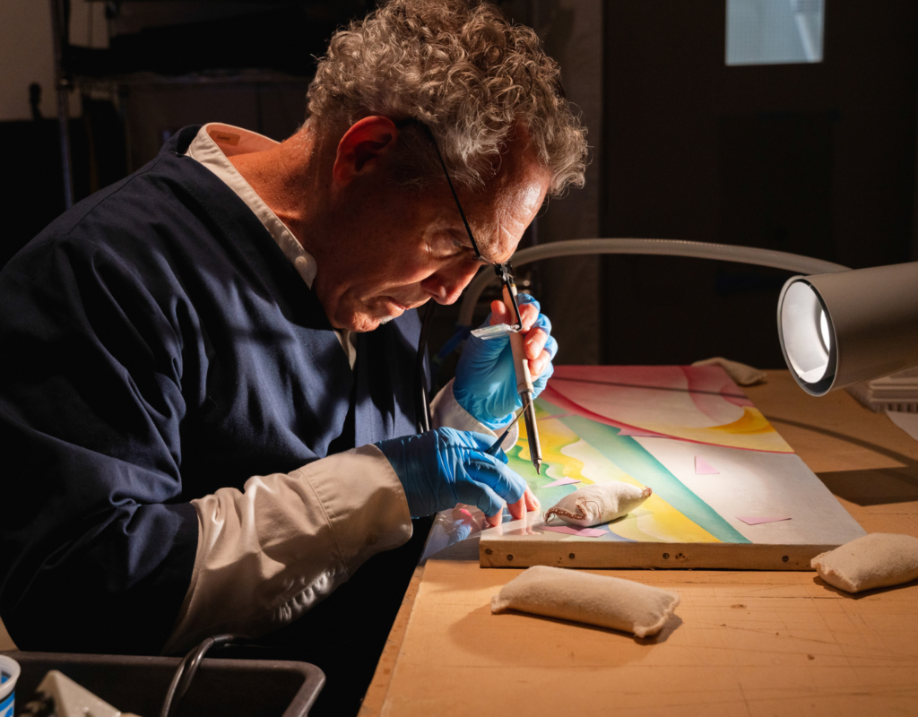 Photograph of a person wearing special eyeglasses and gloves working with specialized tools on a painting set on a table under bright lights.