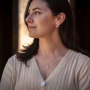 A person with dark hair wearing a silver earring and necklace inspired by the shape of a pelvis.