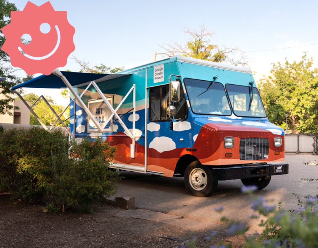 Photograph of the Art to G.O. truck with colorful cloud motifs on it and its awning near some bushes and trees. In the top left corner of the image is a sticker of a pink smiley face, the Risolana logo.