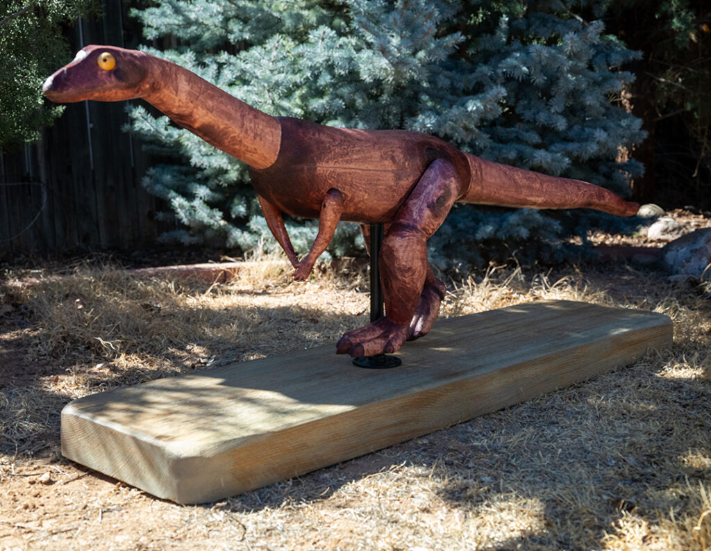Photograph of a wooden model of a small proto-dinosaur, Effigia okeeffeae, on a wooden platform. Behind the sculpture are trees.