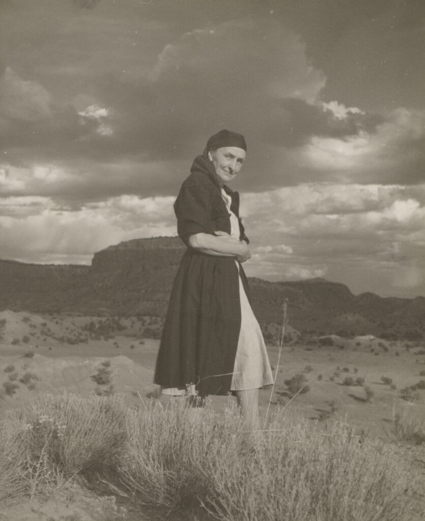 Portrait of Georgia O'Keeffe standing in the New Mexican landscape; profile view of O'Keeffe wearing a black head scarf and black wrap dress with desert grasses in foreground and mountains, sky, and clouds in background.