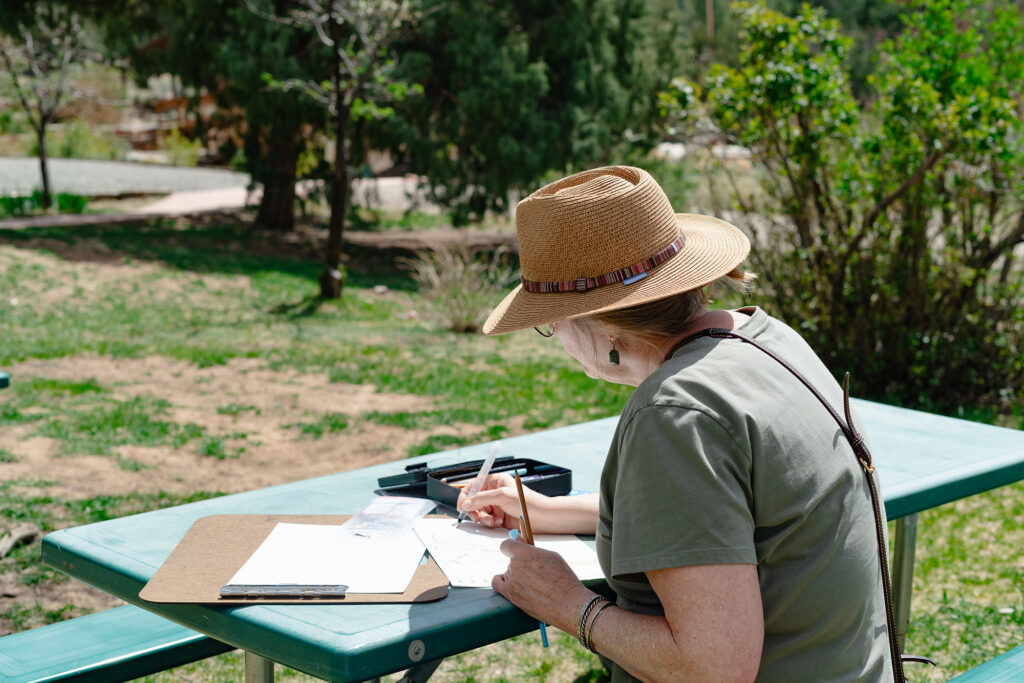 A person with a hat sits at a table sketching and painting. Behind them are green trees and grass.