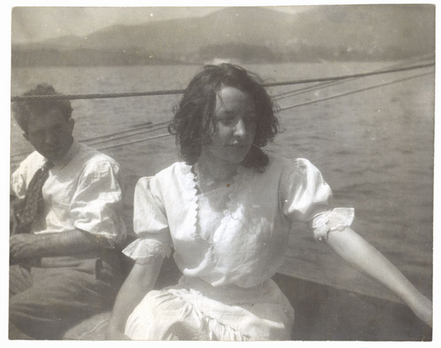 Georgia O'Keeffe wearing a light dress and her hair just below the shoulders sitting in a boat on lake George. To her right is another student from the Art Students League.