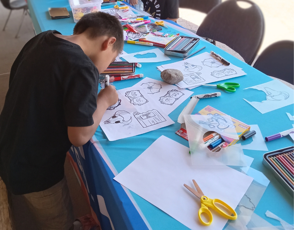 A young person with a dark shirt is color on a white piece of paper. They are bent over the table that is covered with art supplies. The table has a blue table-cloth.