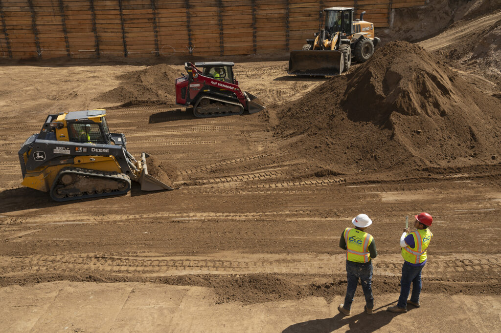 An excavation site with large wooden walls, a dirt bottom, and construction trucks. To the bottom right of the frame two workers stand with hard hats and bright vests. 