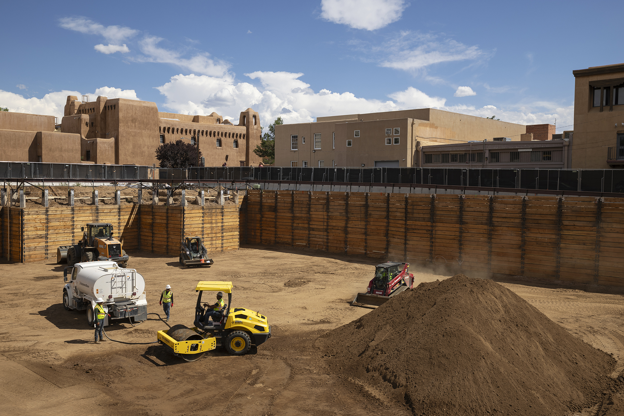 An excavation site with large wooden barriers, a dirt bottom, and construction trucks. Above the site are some adobe buildings and a blue sky.