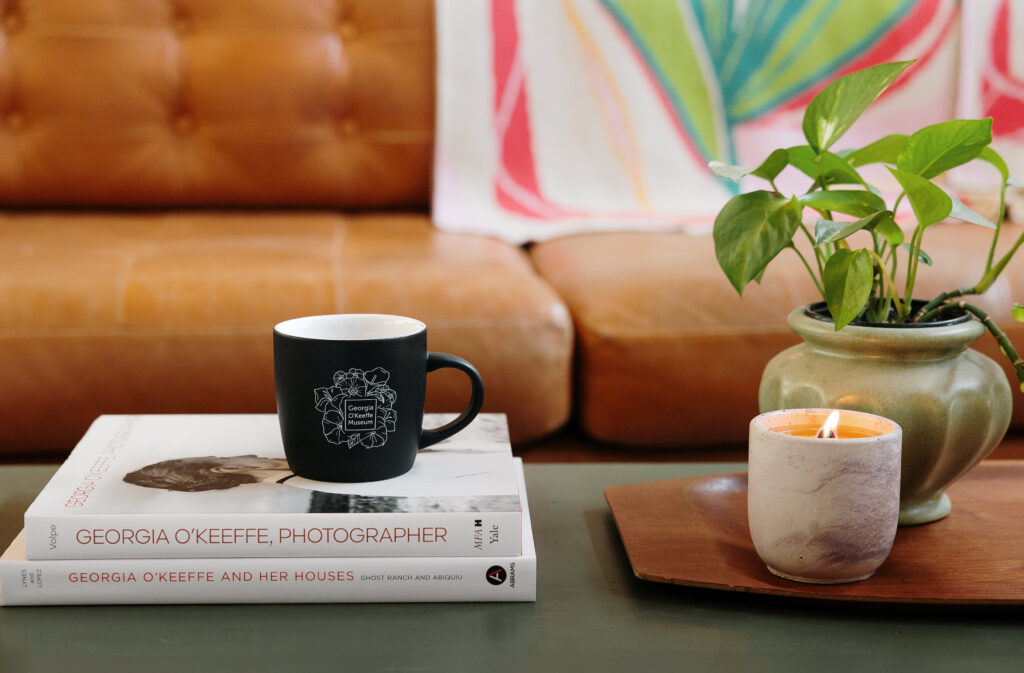 Photograph of a coffee table on which is a stack of books about Georgia O'Keeffe, a black mug with floral designs, a candle with a small flame, and a plant in a pot. Behind is a leather couch on which is draped a blanket in shades of pink and green based abstractly on O'Keeffe's painting of a Pink Tulip.