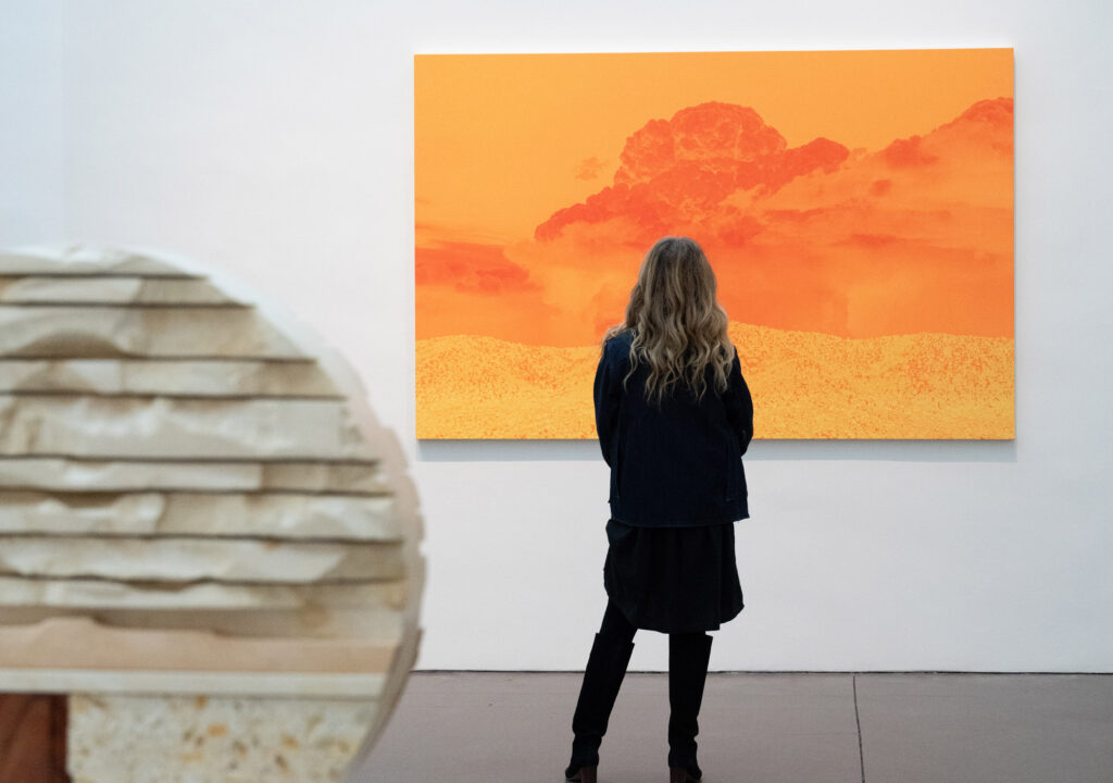 Photograph of a person with long hair from behind standing in front of a large print of clouds over a desert landscape in shades of red and orange. In the foreground is a sculpture in light brown and sand shades.
