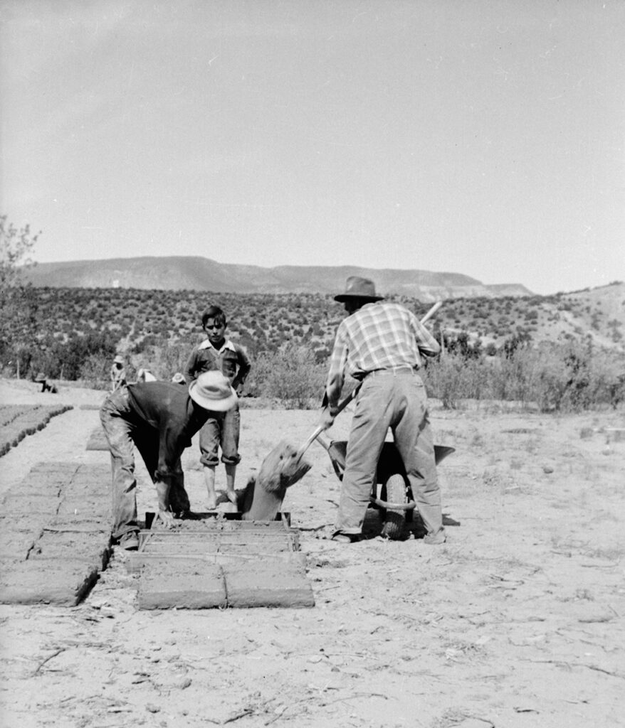 Black and white photograph of two people with a younger child using their hands and a shovel to make adobe bricks from a mud and straw mixture. Behind them is a desert landscape.