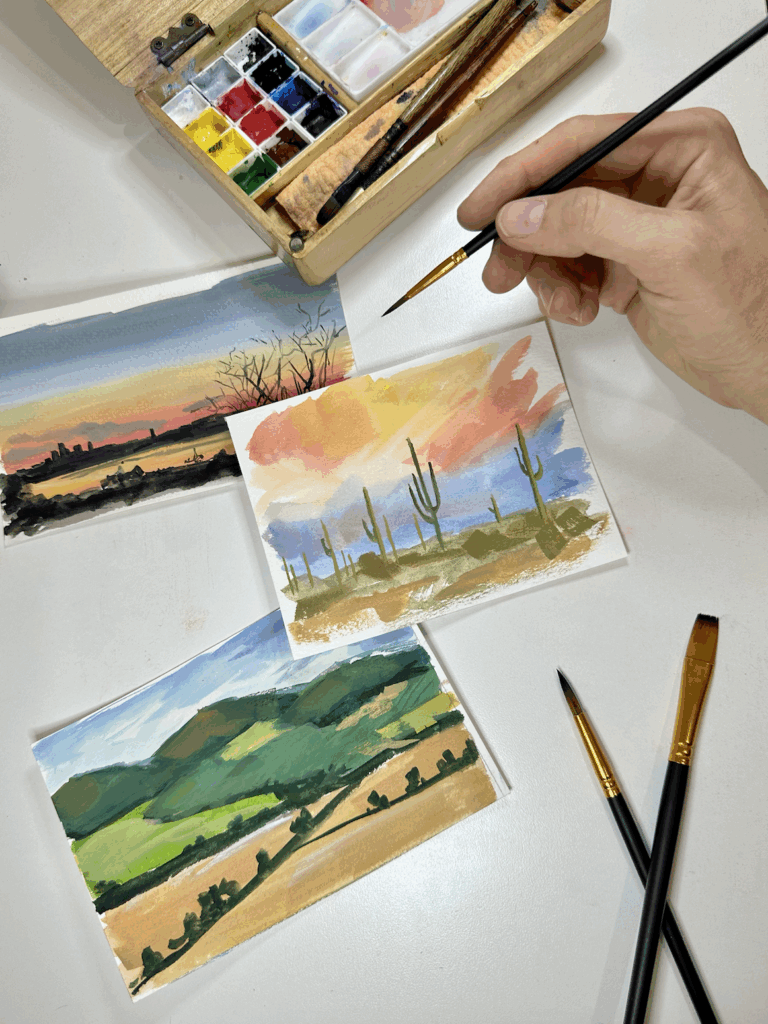 Photograph of three small landscape paintings on a white table. A hand hovers above holding a paintbrush. Nearby are a colorful paint palette and more small paint brushes.