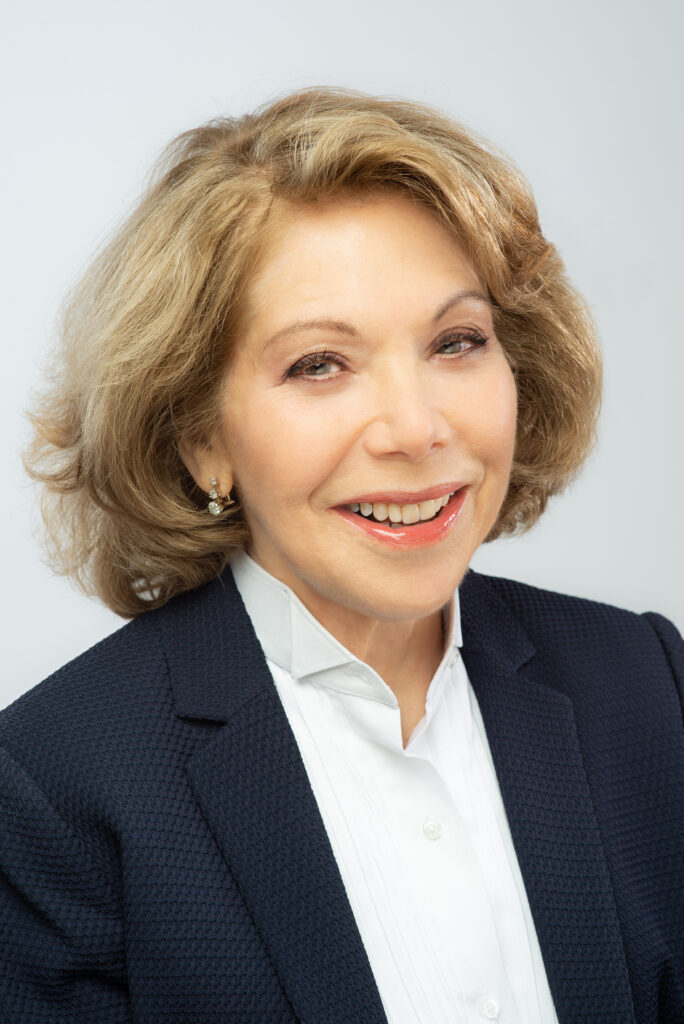 A portrait of composer Victoria Bond wearing a light shirt, dark jacket, and smiling at the camera.