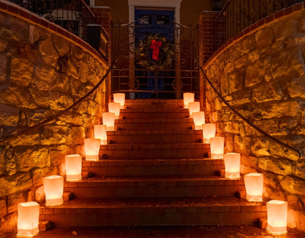 A set of stairs with two rising stone walls on either side lead to a door with a red bow on it. Along the stairs are brown farolitos illuminating the dark.